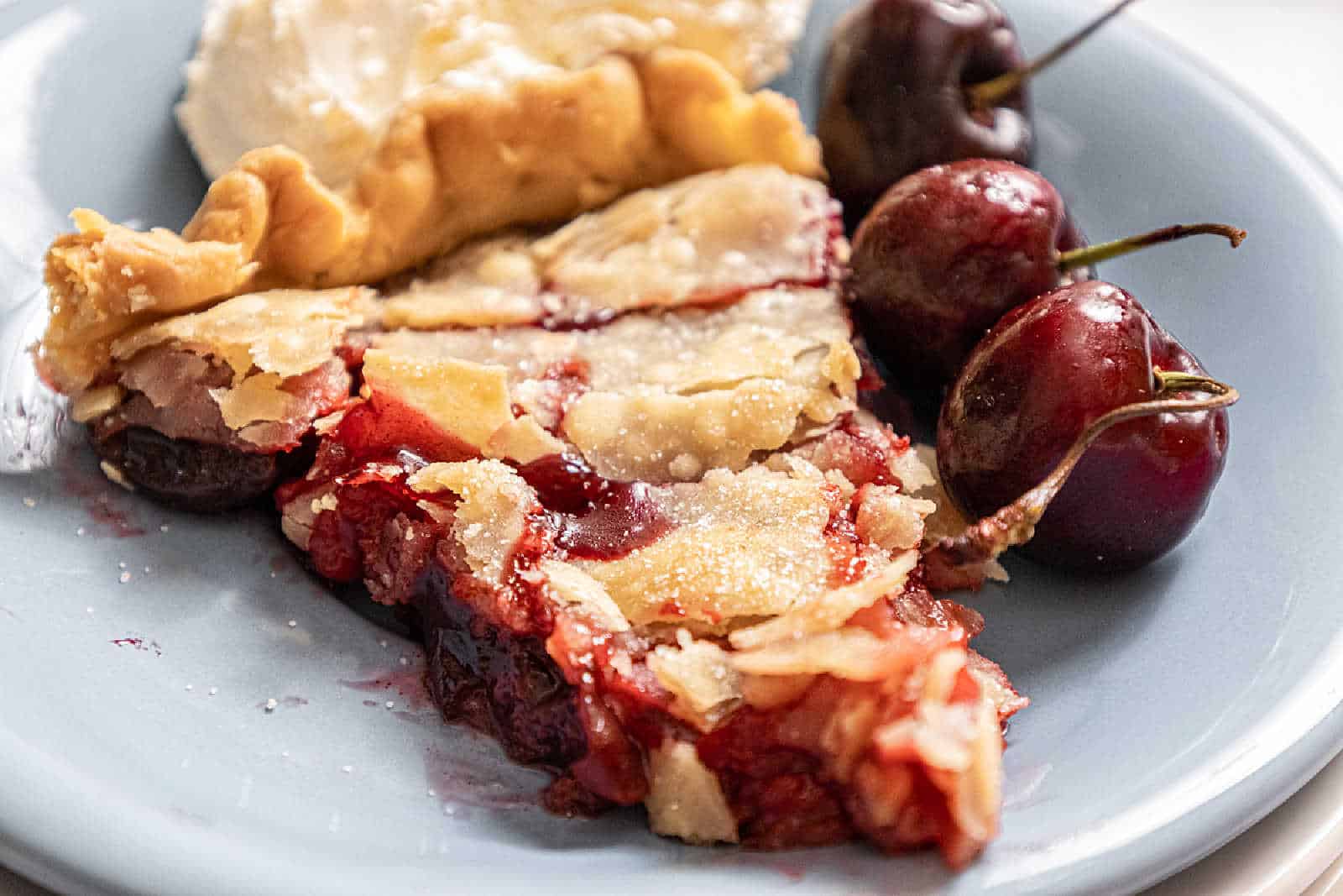 A slice of cherry pie with flaky crust and filling, served on a blue plate with three fresh cherries and a portion of whipped cream.