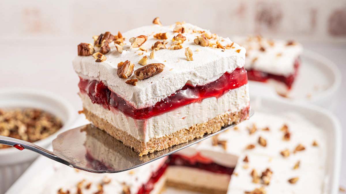 A spatula holds a square slice of layered dessert with a crumb crust, creamy layer, cherry filling, whipped topping, and chopped pecans.
