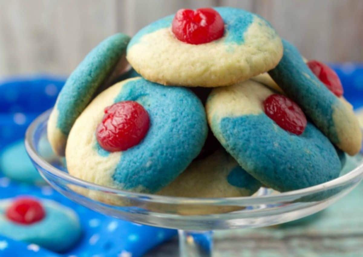 A glass dish holds several sugar cookies with blue coloring and a red cherry on top of each. A blue napkin with white dots is partially visible underneath.