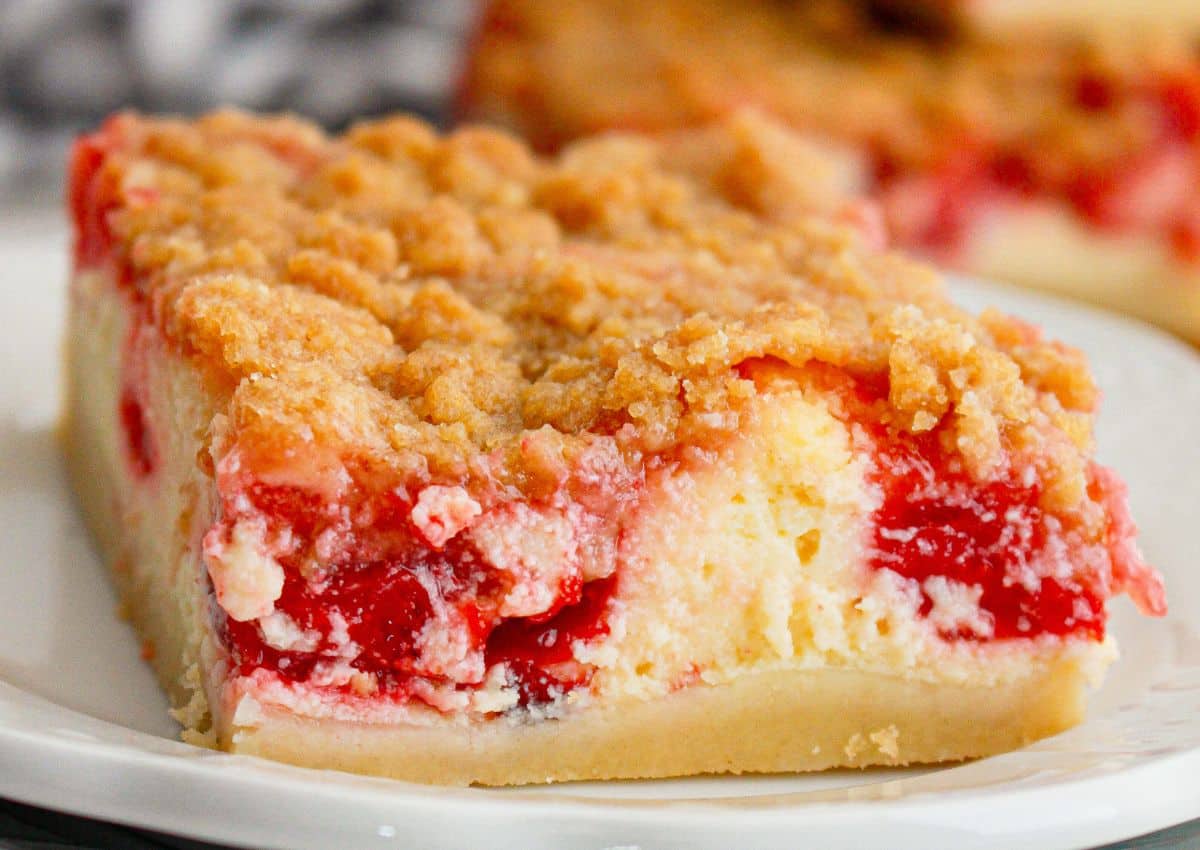 A close-up of a slice of cherry crumb cheesecake bar on a white plate, showing layers of crust, creamy filling, cherry topping, and crumbly streusel.