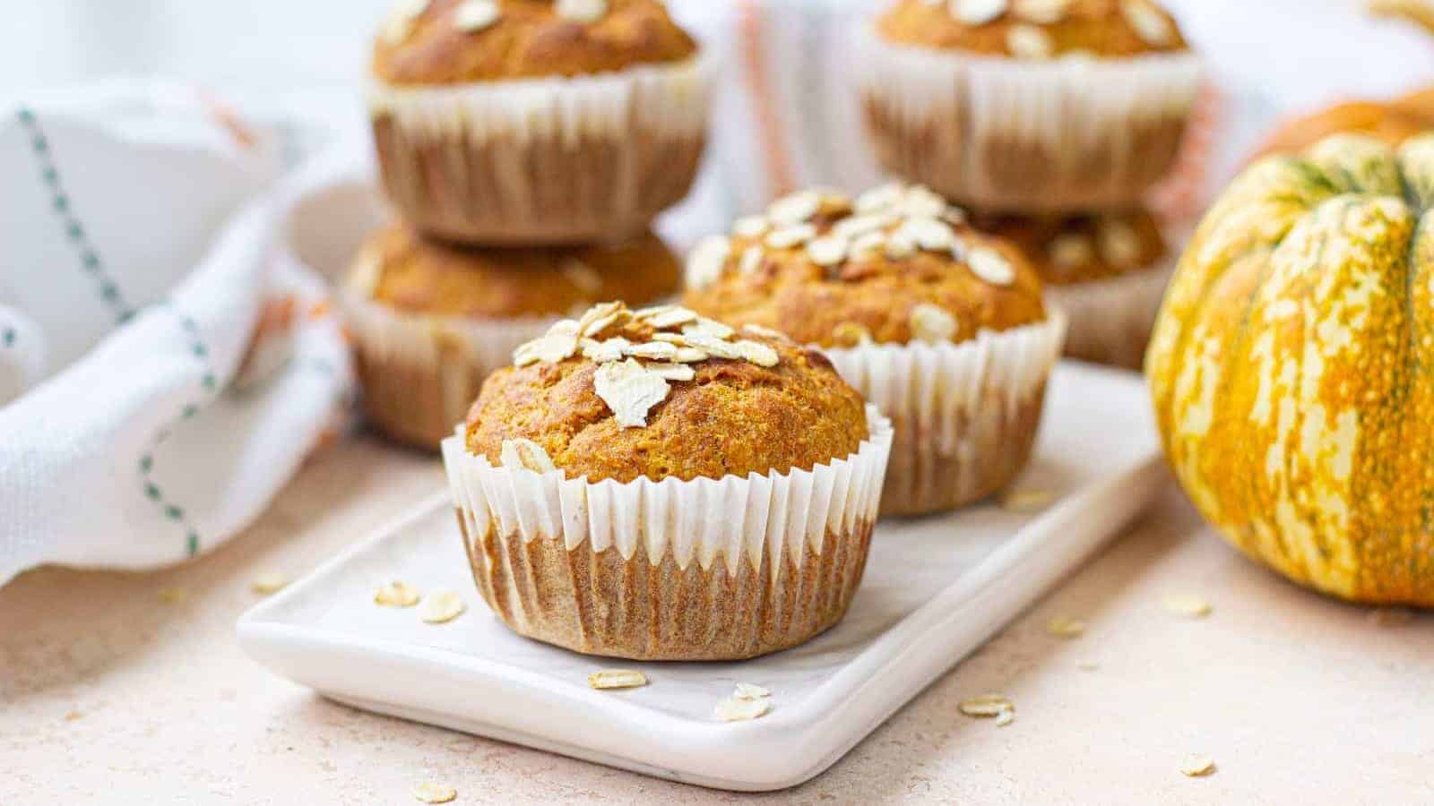 Pumpkin muffins topped with oats are arranged on a white plate, with more muffins and a pumpkin in the background.