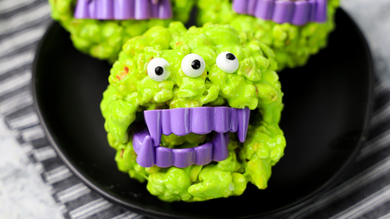 A green, monster-themed treat with candy eyes and purple plastic fangs sits on a black plate.