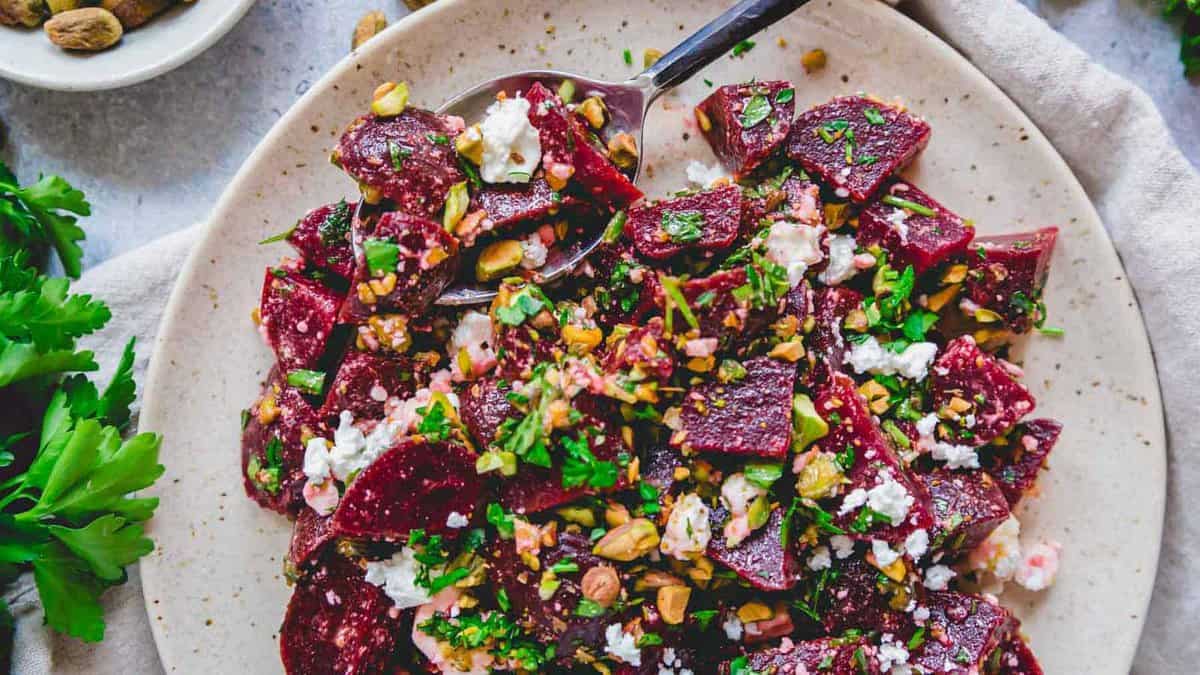 A plate of beet salad topped with crumbled cheese, chopped nuts, and fresh herbs, with a serving spoon on the side.