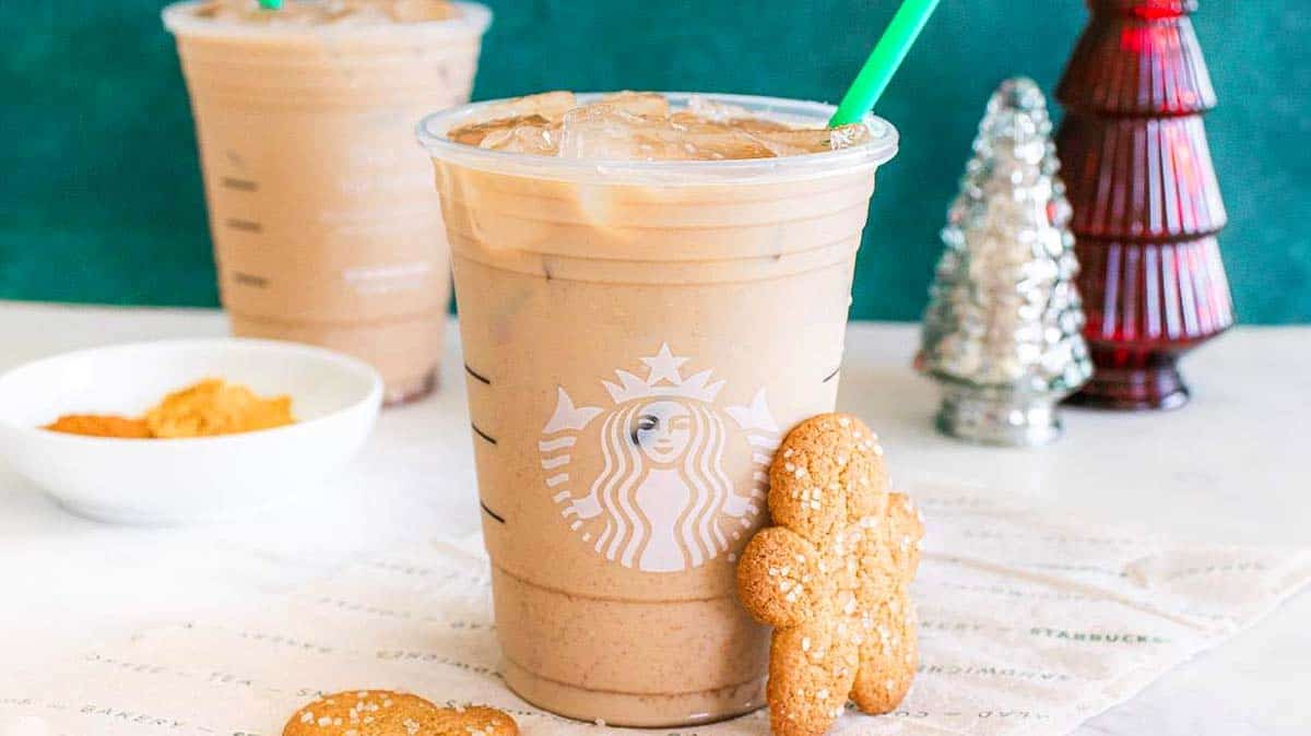 An iced coffee in a clear Starbucks cup with a green straw, next to a gingerbread cookie and festive holiday decorations in the background.