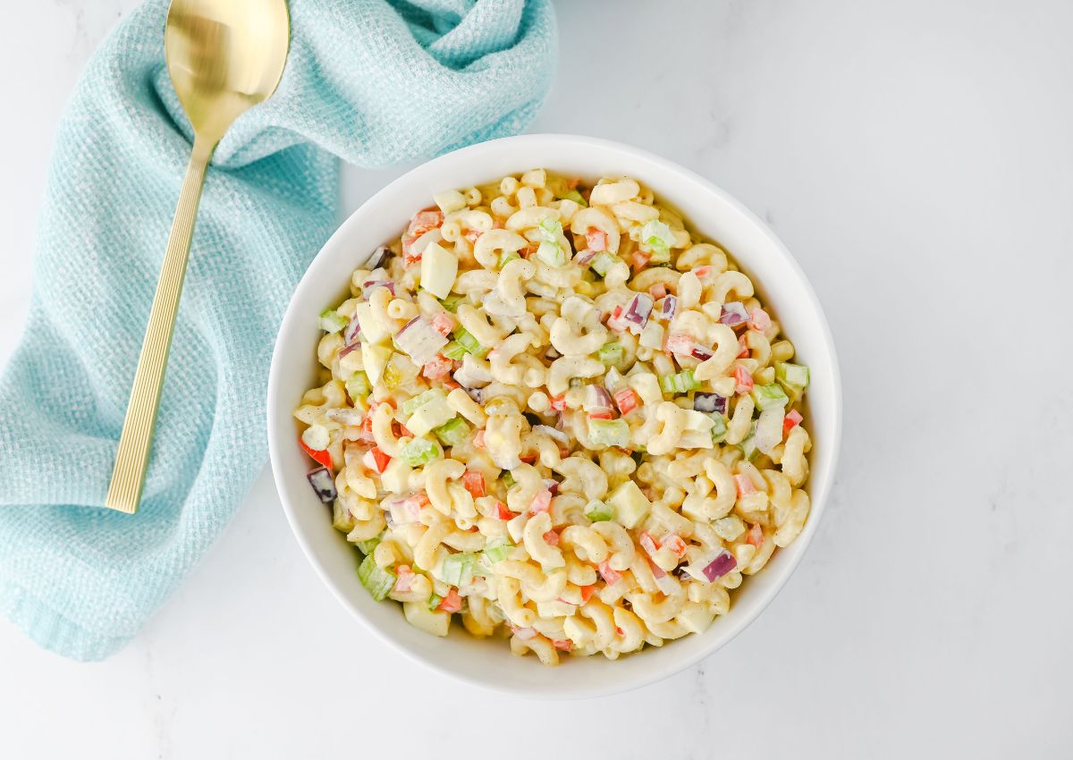 A white bowl filled with macaroni salad containing diced vegetables sits next to a gold spoon and a light blue cloth on a white surface.