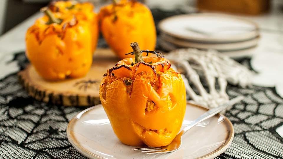 A baked yellow bell pepper carved with a jack-o-lantern face sits on a plate with a fork; more stuffed peppers are in the background on a wooden tray.