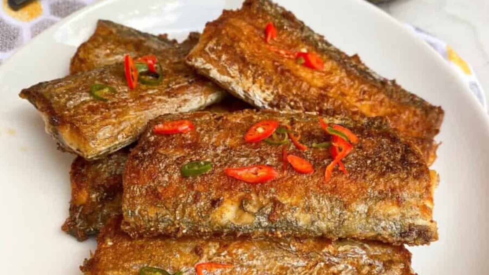 Several pieces of grilled or fried fish fillets garnished with sliced red and green chili peppers, served on a white plate.