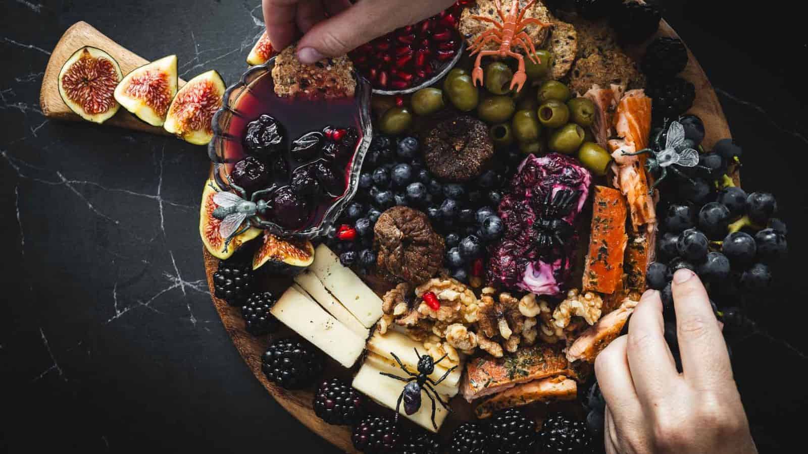A person arranges items on a wooden charcuterie board with cheese, figs, berries, nuts, olives, crackers, and pomegranate seeds on a dark surface.