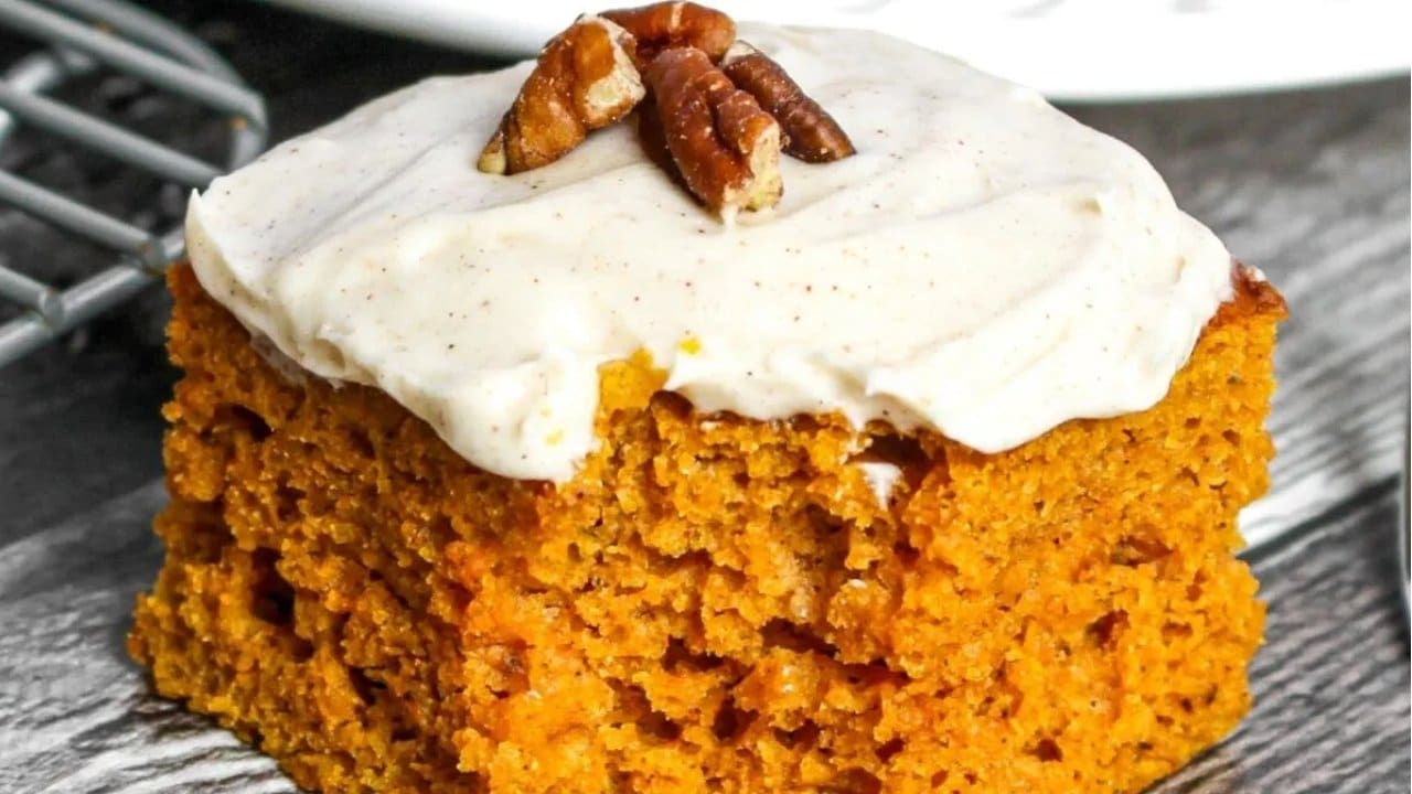 A slice of pumpkin cake with white frosting topped with pecan halves, placed on a dark surface.