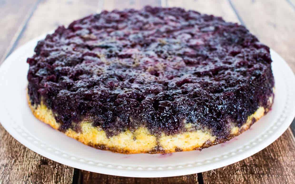 A round blueberry upside-down cake sits on a white plate, displaying a dense layer of baked blueberries on top.