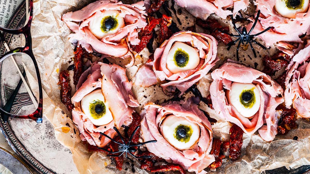 Slices of ham arranged with mozzarella and olives to resemble eyes, surrounded by sun-dried tomatoes and plastic spider decorations on parchment paper.