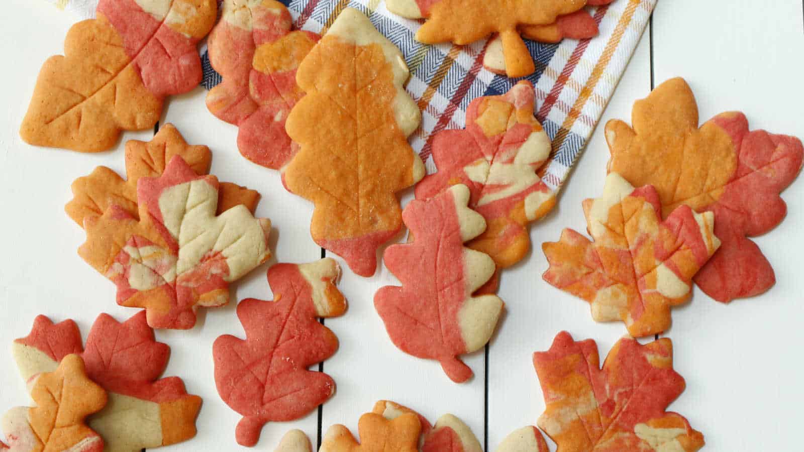Assorted leaf-shaped cookies in autumn colors are arranged on a white surface next to a plaid cloth.