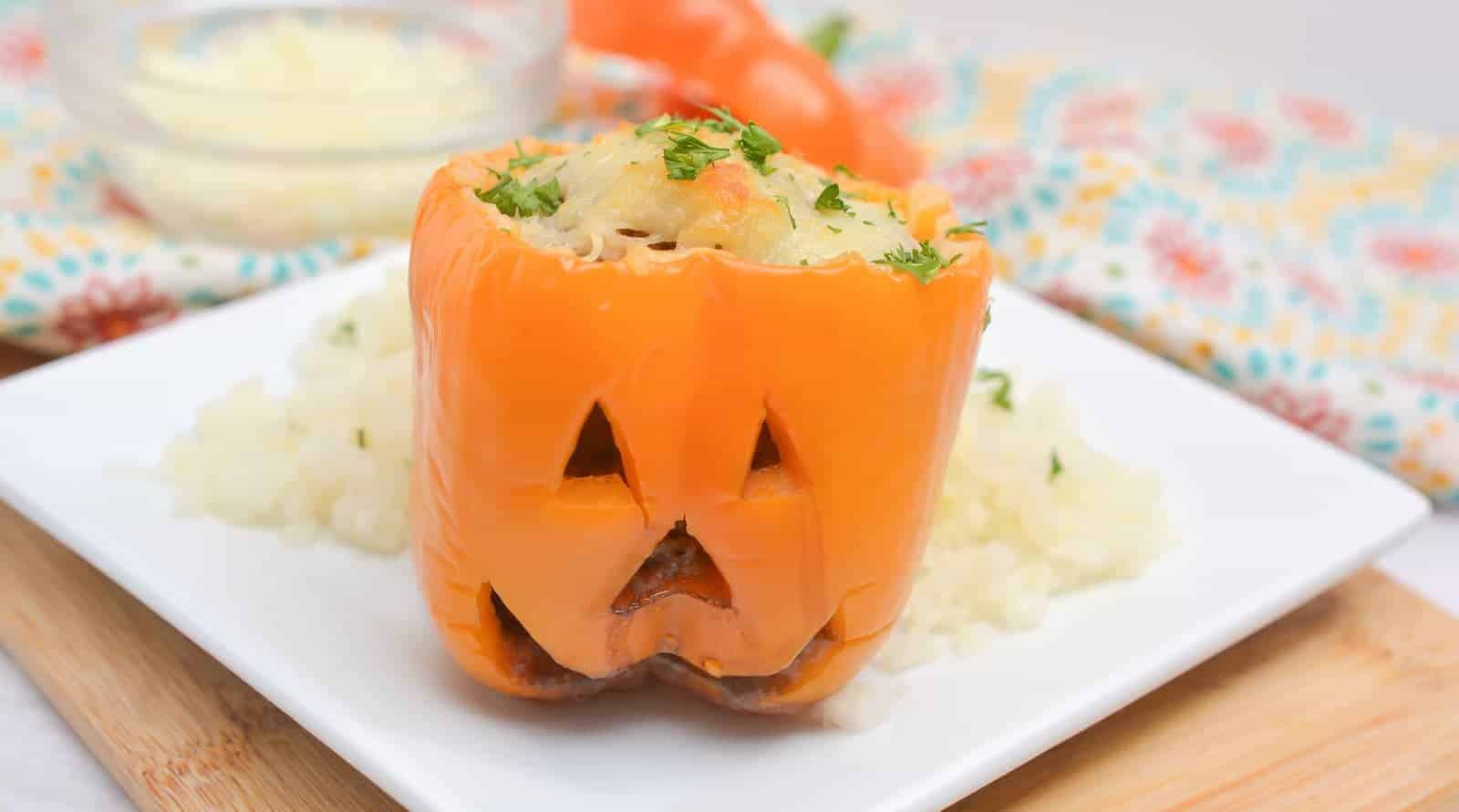 An orange bell pepper carved with a jack-o-lantern face, stuffed with filling, served on a white plate with mashed potatoes in the background.