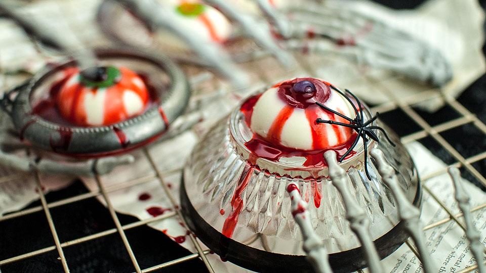 Glass bowls hold white and red dome-shaped desserts resembling eyeballs, topped with fake blood and plastic spiders, on a cooling rack with newspaper beneath.