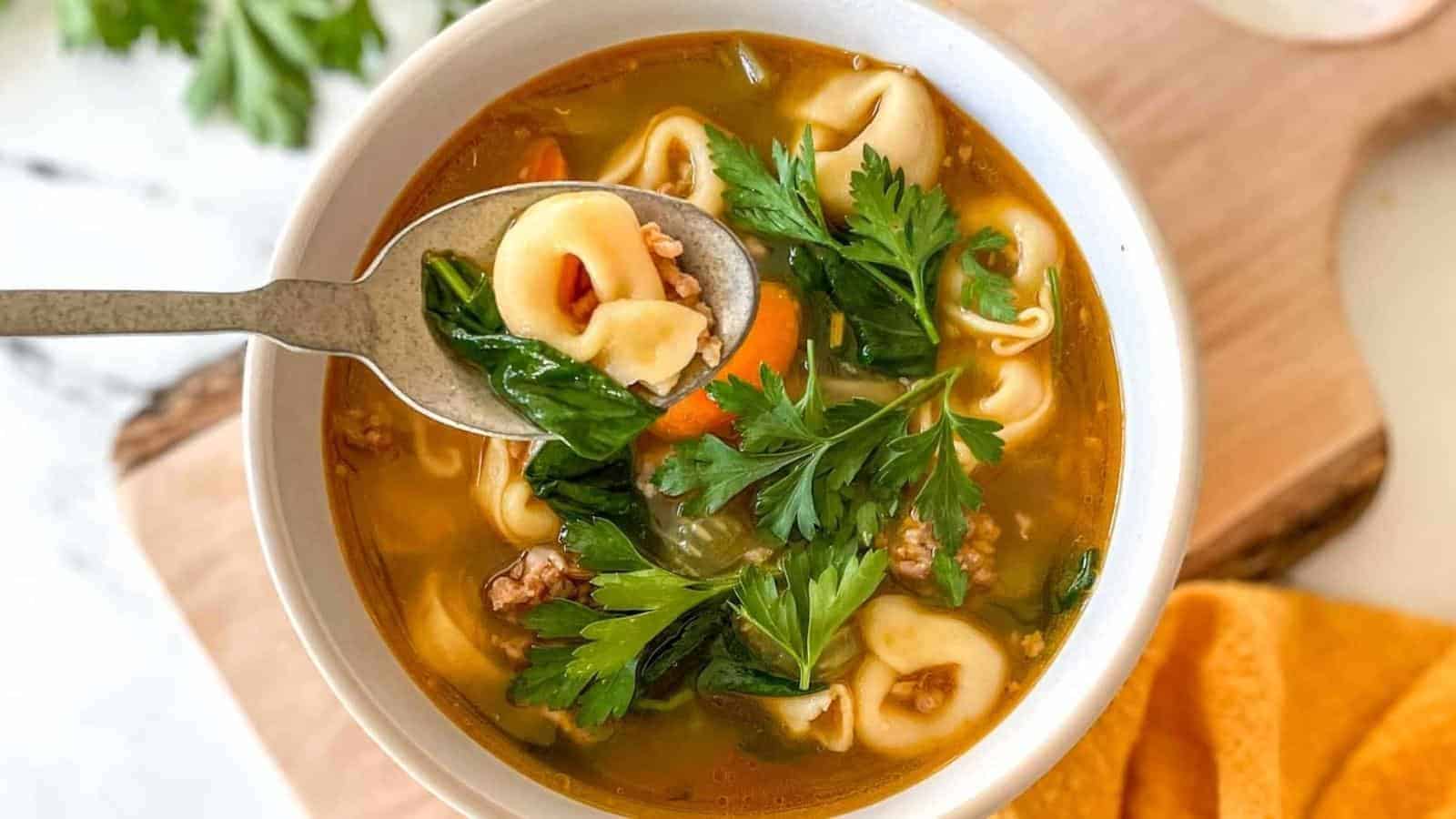 A bowl of tortellini soup with vegetables, ground meat, and fresh parsley, with a spoon lifting some tortellini and herbs.