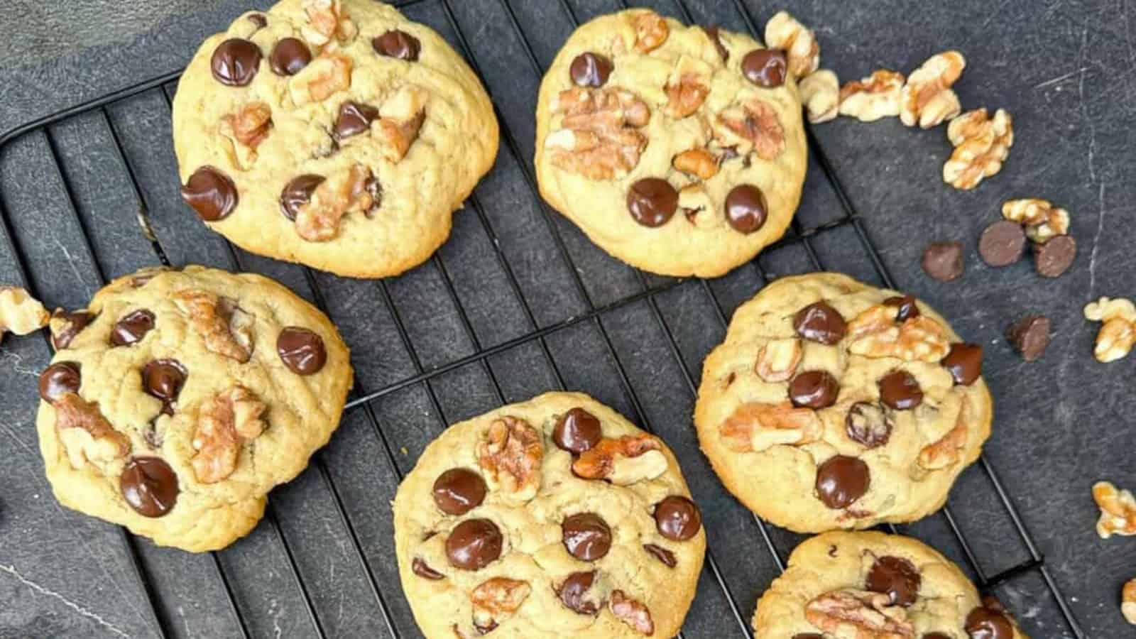 Six chocolate chip and walnut cookies are cooling on a black wire rack, with extra walnut pieces and chocolate chips scattered nearby.