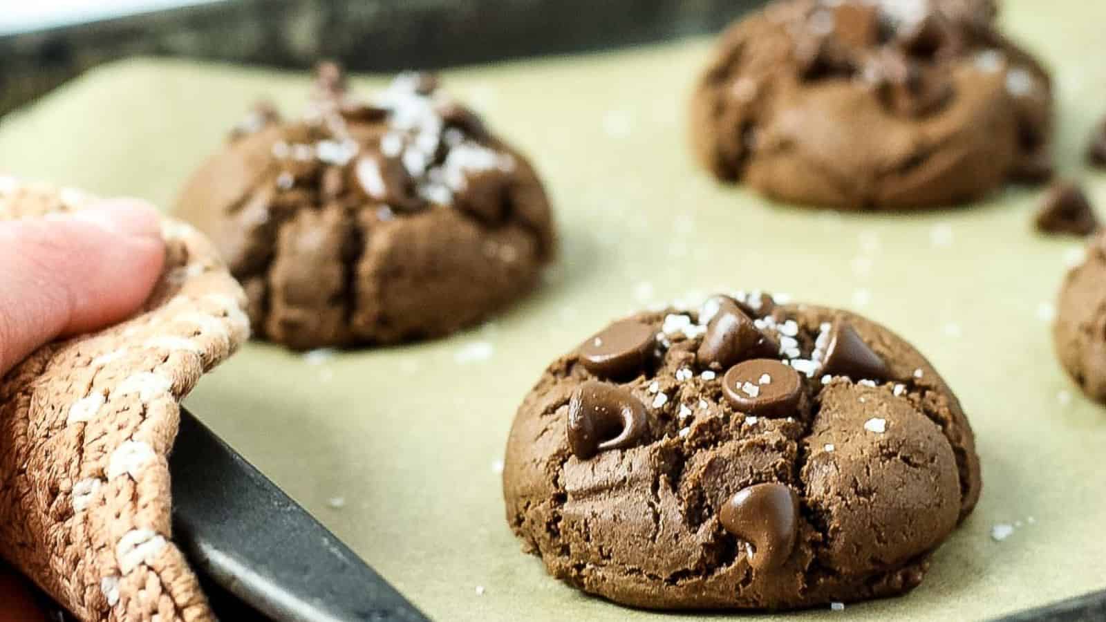 Close-up of freshly baked chocolate cookies with chocolate chips and sea salt on parchment paper, with a hand holding a baking tray.