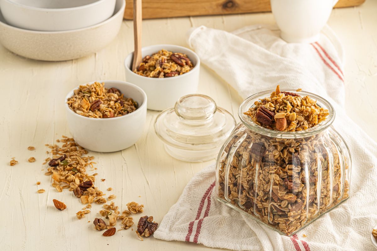 A glass jar filled with granola sits on a towel, with granola spilled around and two white bowls of granola nearby on a light-colored table.