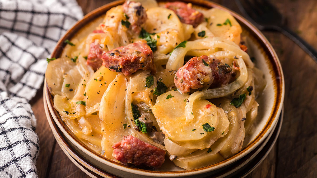A bowl of sliced potatoes, onions, and sausage in a creamy sauce, garnished with chopped herbs, sits on a wooden table beside a checkered cloth.