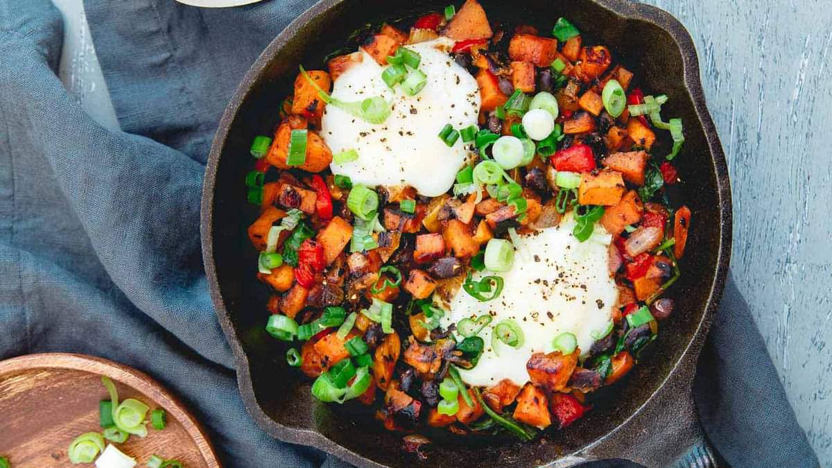 A cast iron skillet filled with sweet potato hash, diced vegetables, two eggs, and topped with chopped green onions.