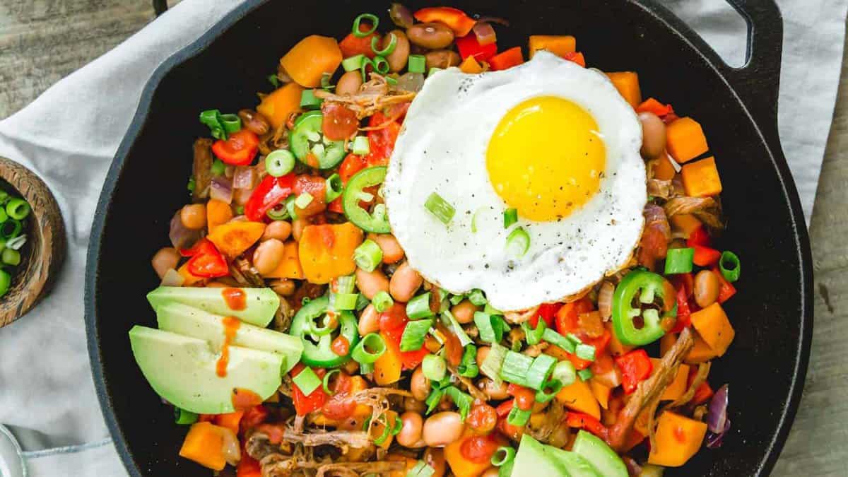 A skillet filled with sweet potatoes, beans, peppers, pulled meat, sliced avocado, green onions, jalapeños, and a sunny-side-up egg on top.