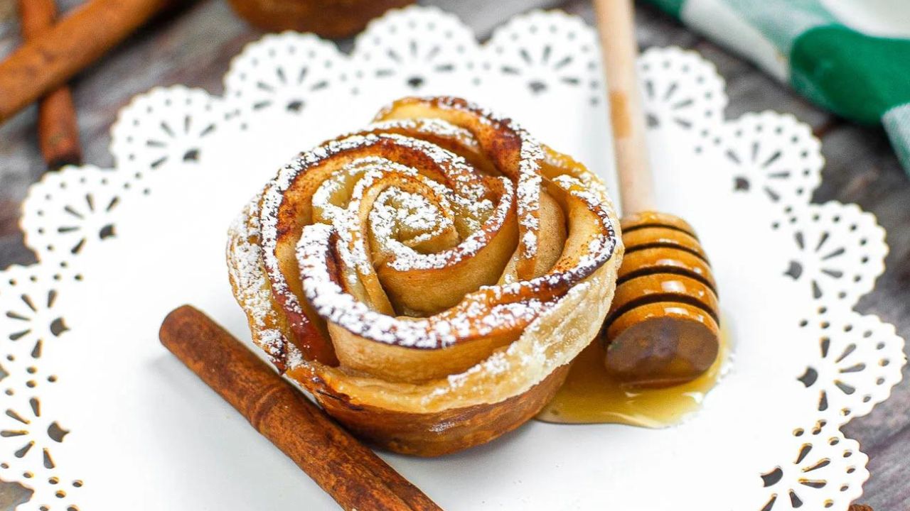 A pastry shaped like a rose, dusted with powdered sugar, is on a white plate with a cinnamon stick and a honey dipper drizzled with honey.