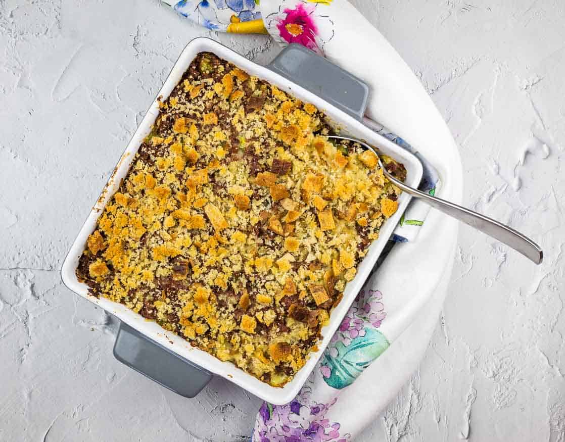 A square baking dish filled with a baked dessert topped with crumbly golden brown streusel, set on a white floral-patterned kitchen towel with a spoon inside.