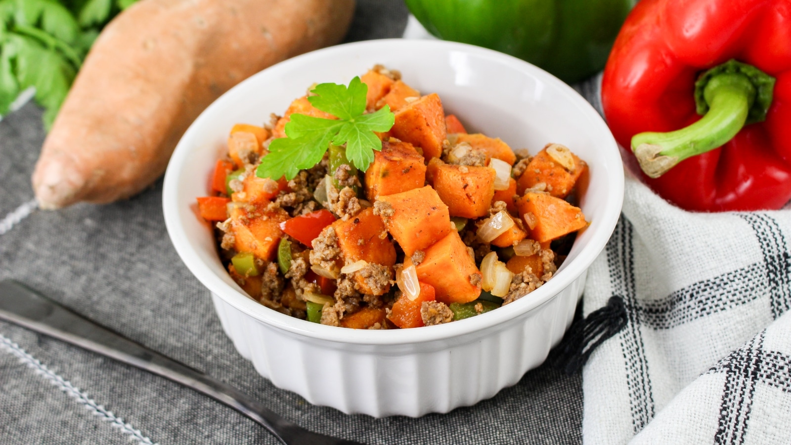 A white bowl filled with a cooked mixture of ground meat, sweet potatoes, red bell peppers, onions, and herbs, garnished with parsley, placed on a cloth with vegetables beside it.