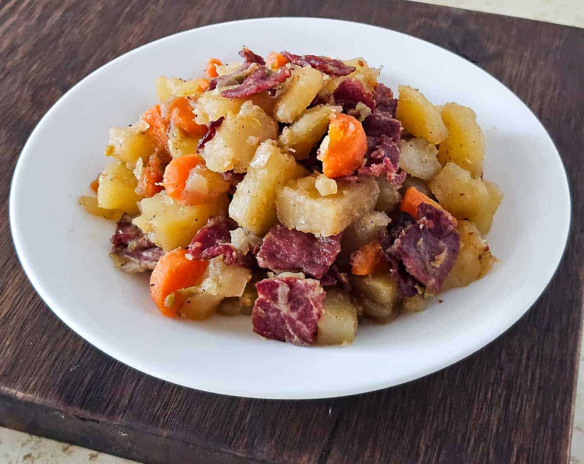 A white plate filled with cooked diced potatoes, carrots, and pieces of corned beef, placed on a dark wooden surface.