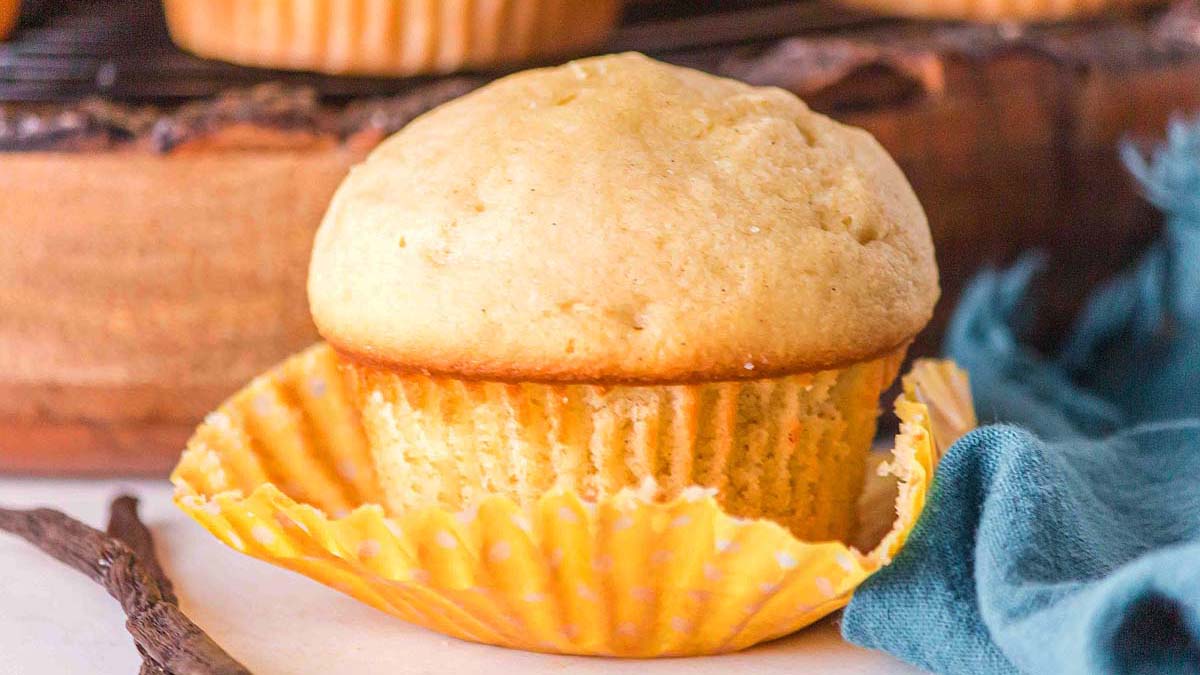 A plain vanilla muffin in a yellow polka-dotted paper liner sits partially unwrapped on a white surface, with a blue cloth and wooden tray in the background.