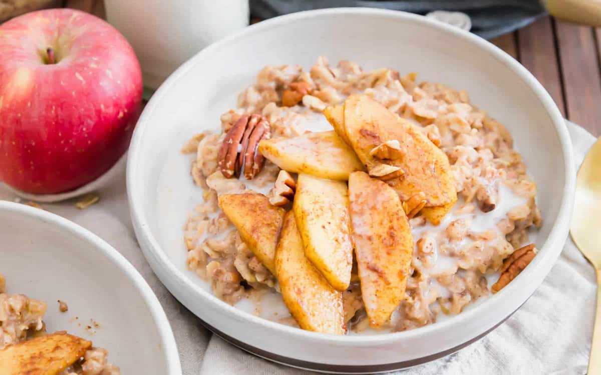 A bowl of oatmeal topped with caramelized apple slices, pecans, and a drizzle of milk, with a red apple visible in the background.