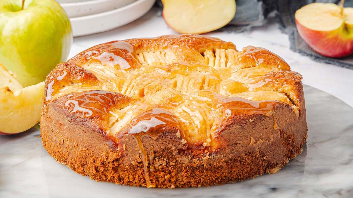 A round apple cake with a glossy glaze sits on a marble surface, surrounded by fresh apples and apple slices.