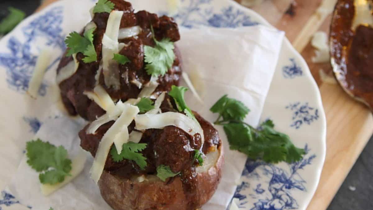 Two baked potatoes topped with shredded cheese, chunks of meat in sauce, and fresh cilantro are served on a white plate with a blue floral pattern.