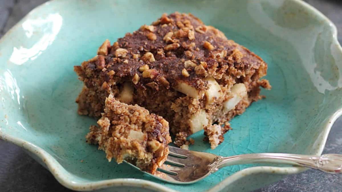 A square slice of apple walnut cake with a crumbly topping sits on a teal plate; a fork rests in front, holding a bite of the cake.