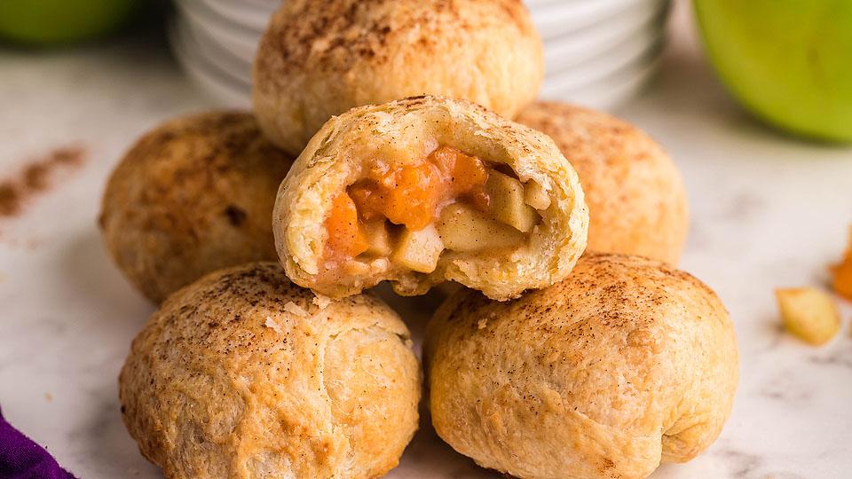 A stack of baked pastry balls, one cut open to reveal a filling of diced apples and sauce, resting on a marble surface.