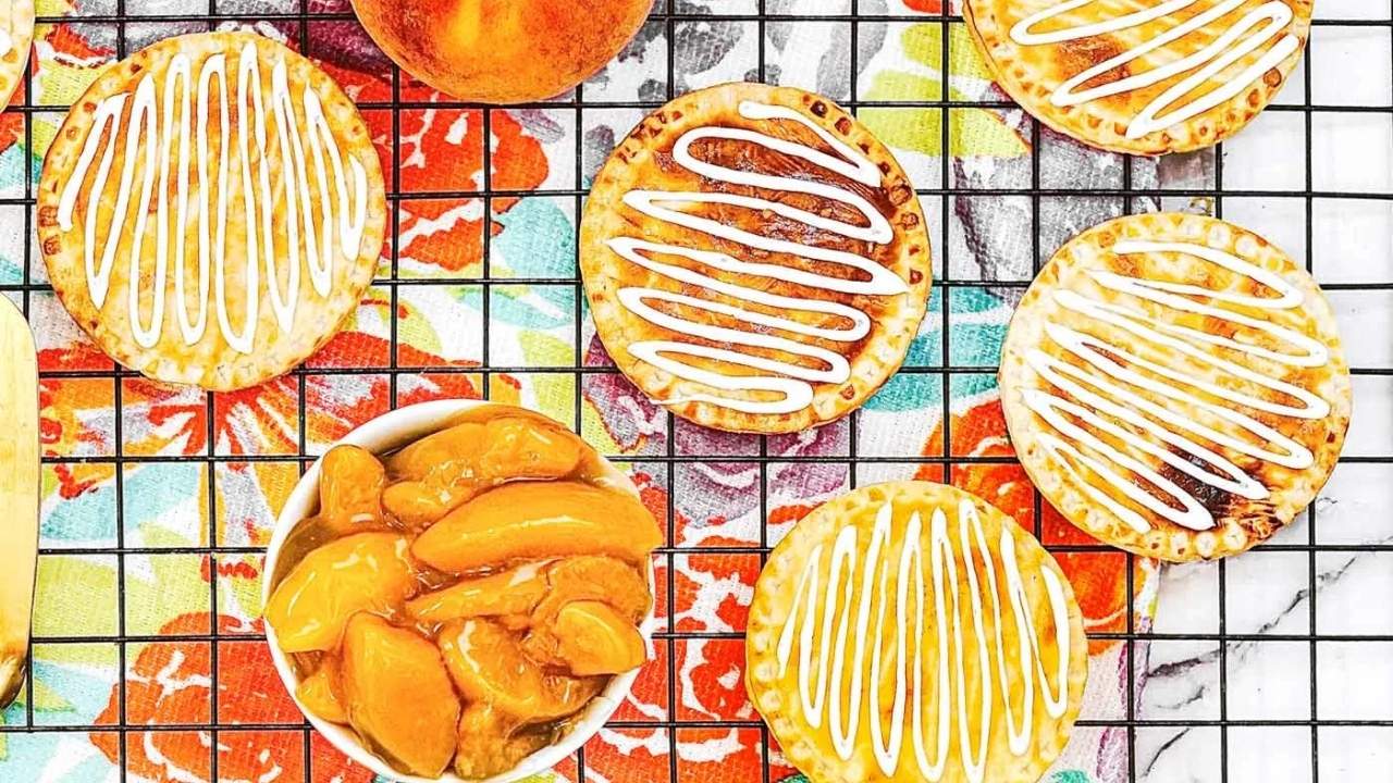 Round pastries with white icing are cooling on a wire rack next to a bowl of sliced peaches and a whole peach, all on a colorful floral cloth.