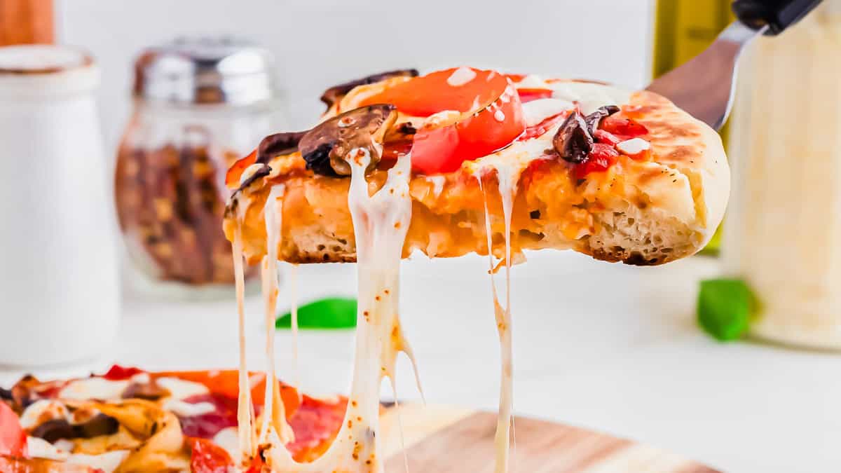 A close-up of a slice of pizza with melted cheese being lifted from a whole pizza, showing toppings like tomatoes and mushrooms.