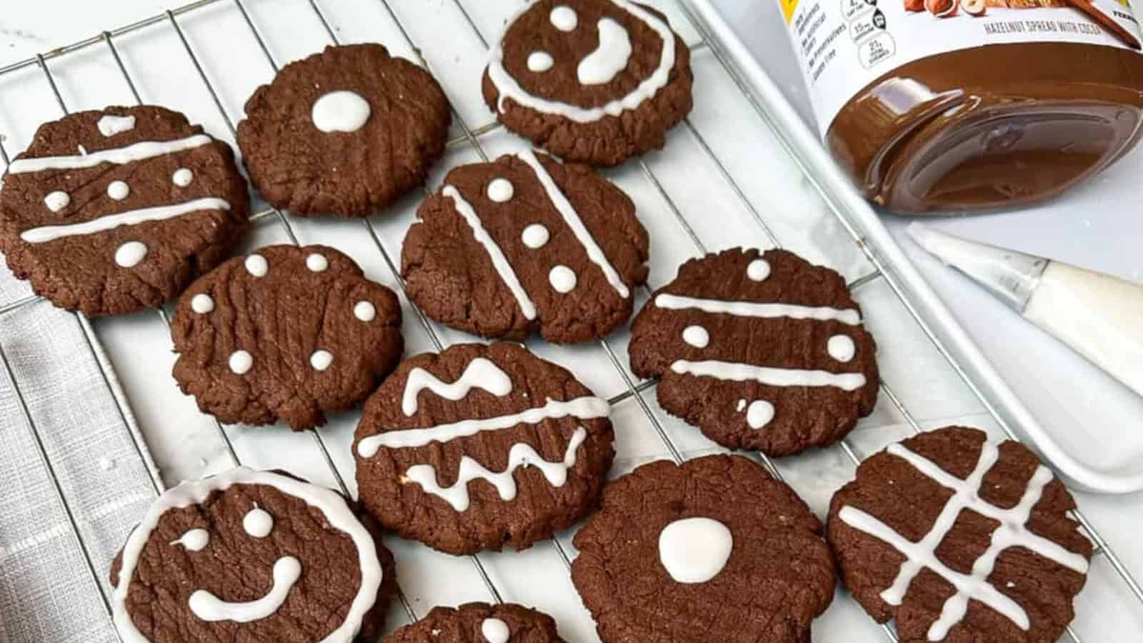 Chocolate cookies with white icing decorations are arranged on a cooling rack next to a jar of chocolate spread and a butter knife.