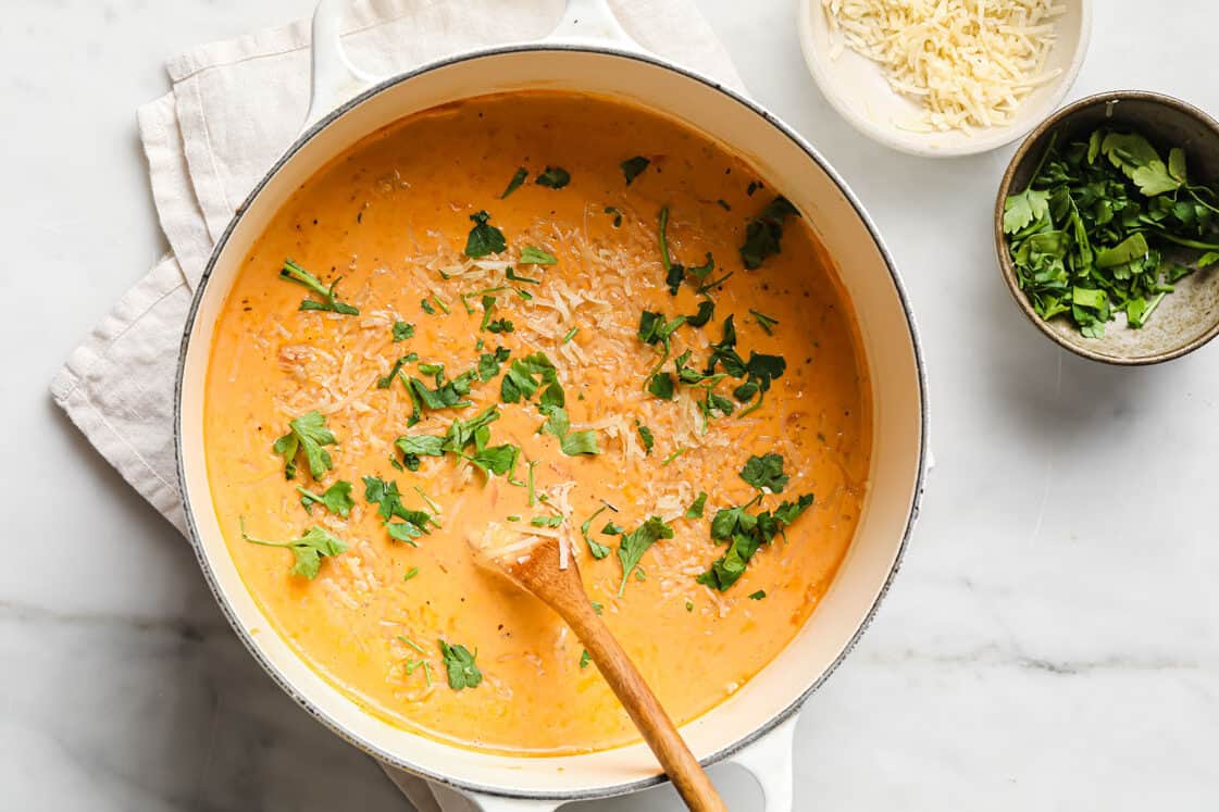 A pot of creamy tomato soup garnished with chopped parsley and shredded cheese, with a wooden spoon resting inside and bowls of ingredients nearby.