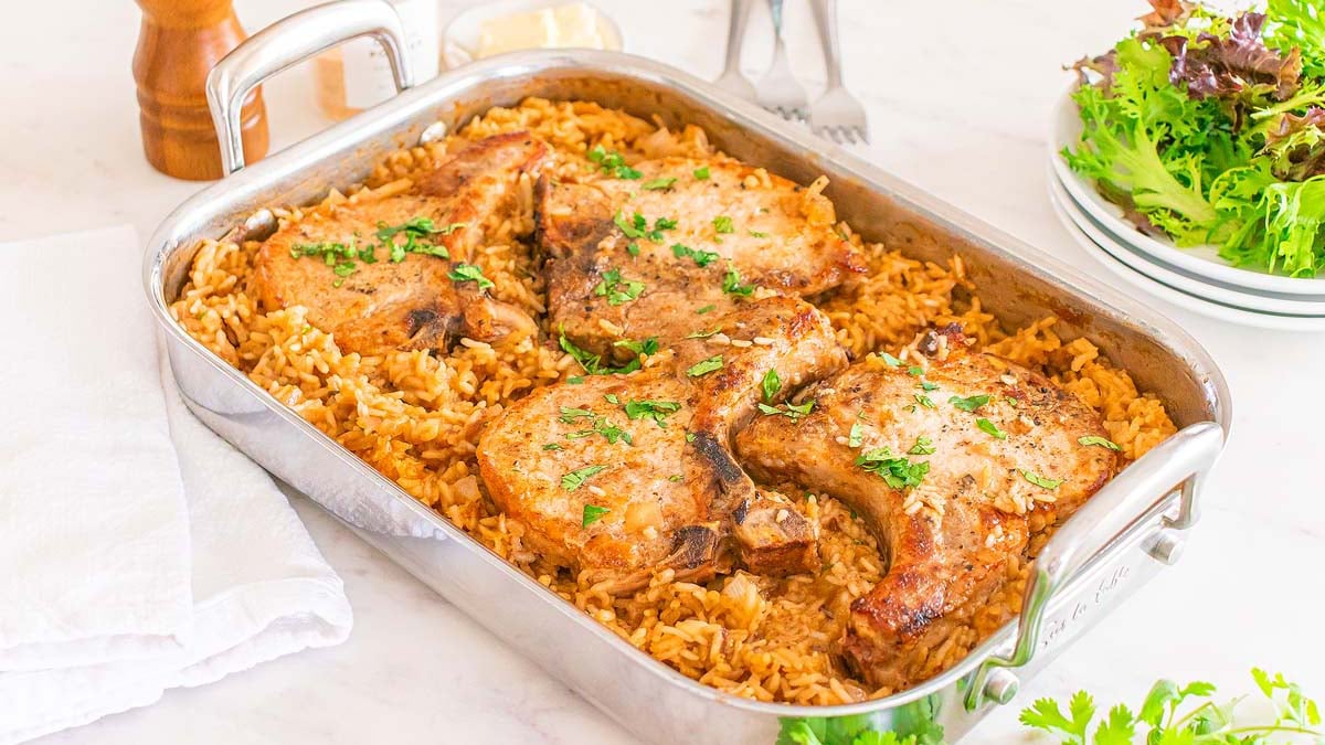 Baked pork chops with herbs served on a bed of seasoned rice in a rectangular metal roasting pan, with a side salad and plates in the background.