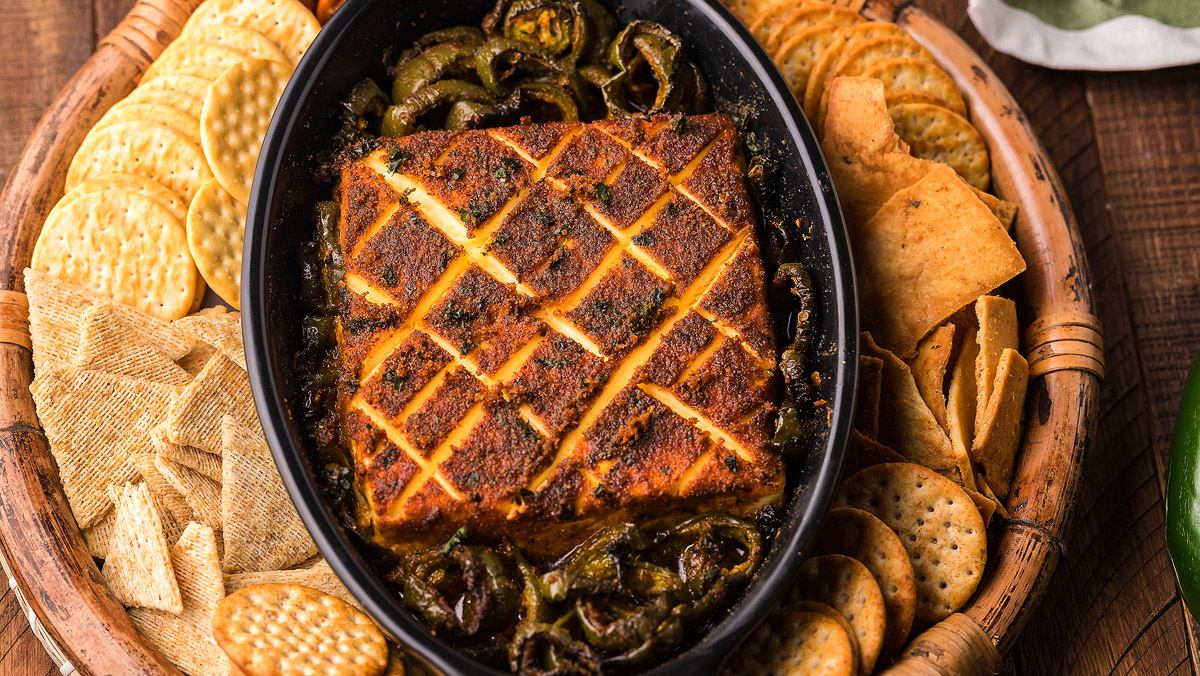 A seasoned, baked block of cheese with a crisscross pattern sits in a black dish, surrounded by assorted crackers and pita chips on a wooden tray.