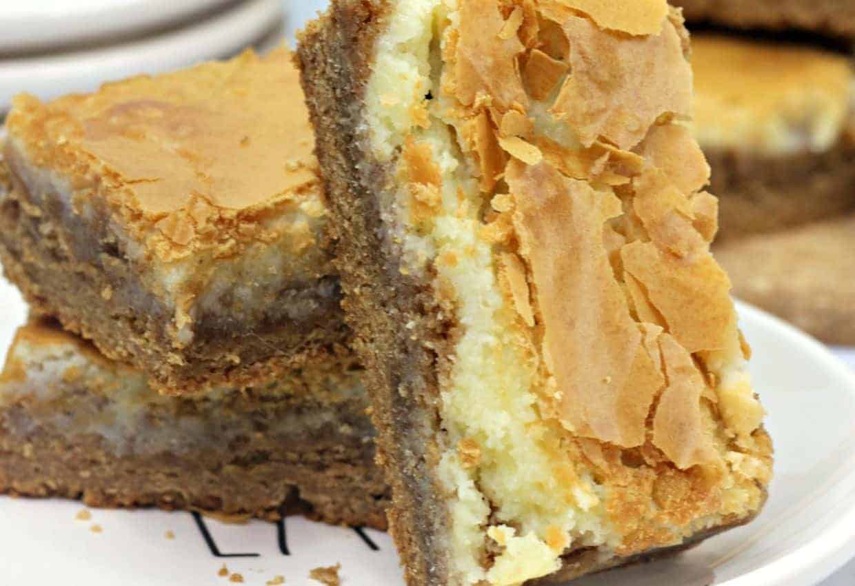 Two gooey butter cake bars with a golden, flaky top and creamy filling are stacked on a white plate.