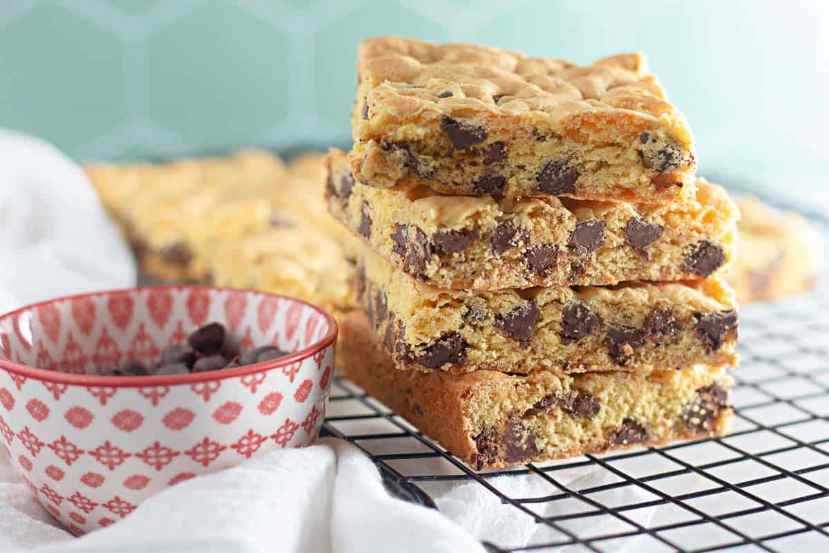 Four chocolate chip cookie bars are stacked on a cooling rack next to a patterned bowl containing chocolate chips.