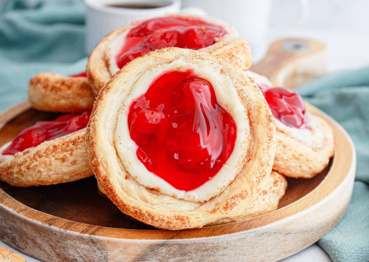 A stack of cherry cheese danishes on a wooden plate with a cup of coffee in the background.