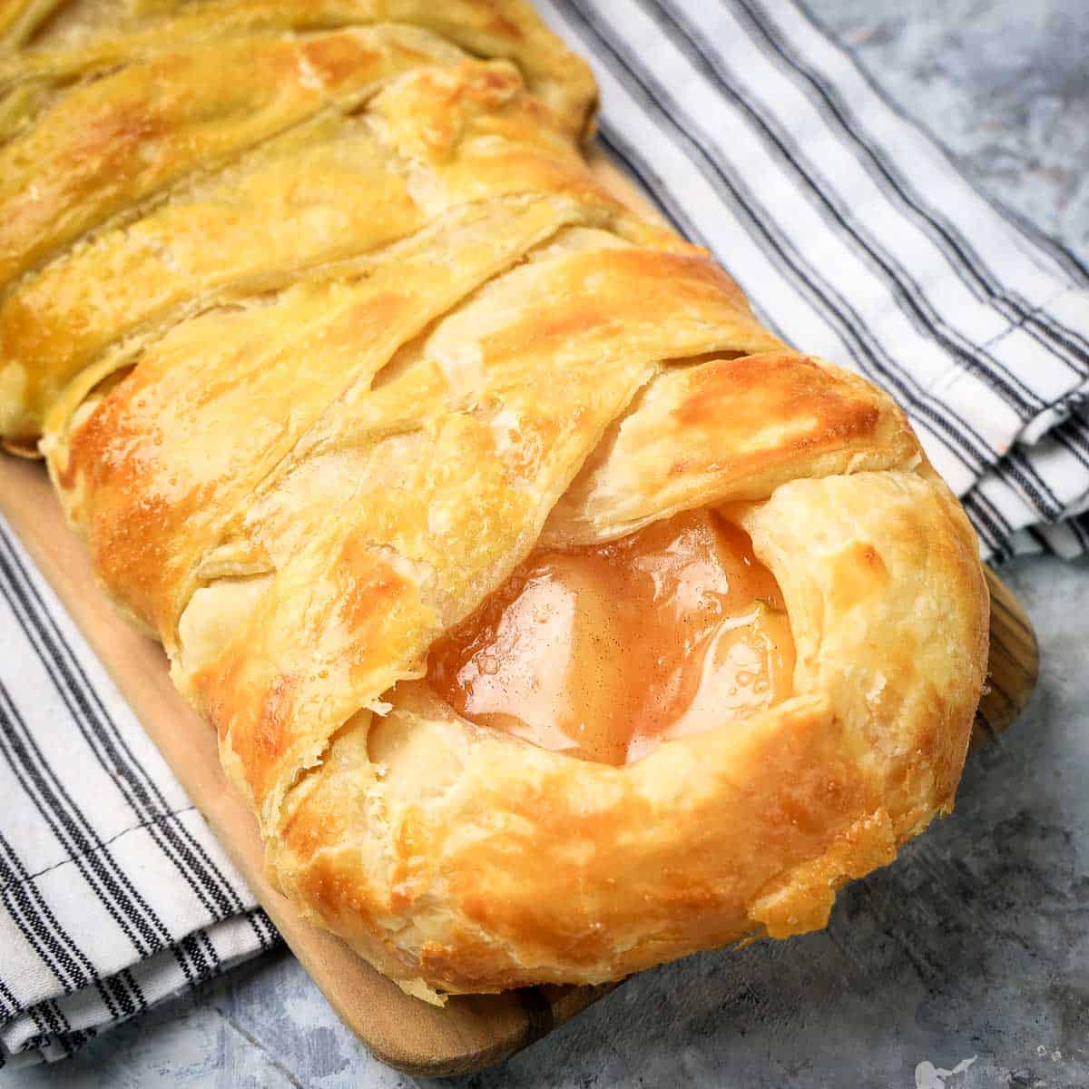 Golden brown braided pastry with a visible apple filling on a wooden board, set on a striped kitchen towel.