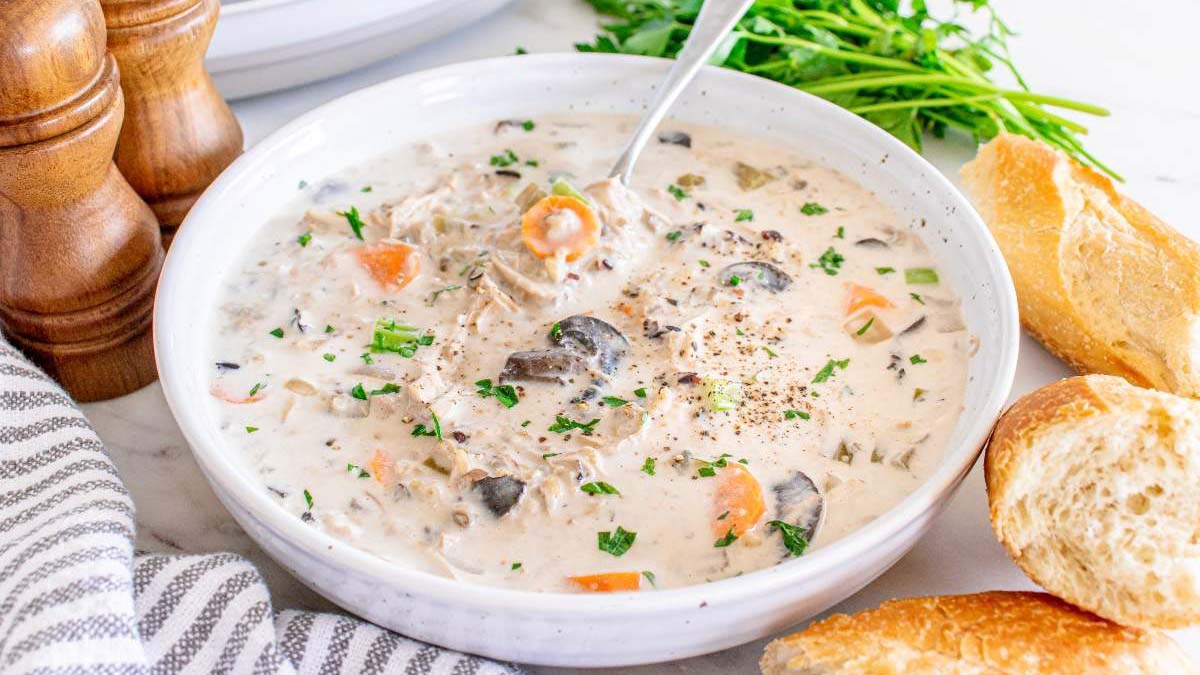 A bowl of creamy chicken and vegetable soup garnished with herbs, served with bread slices on the side.