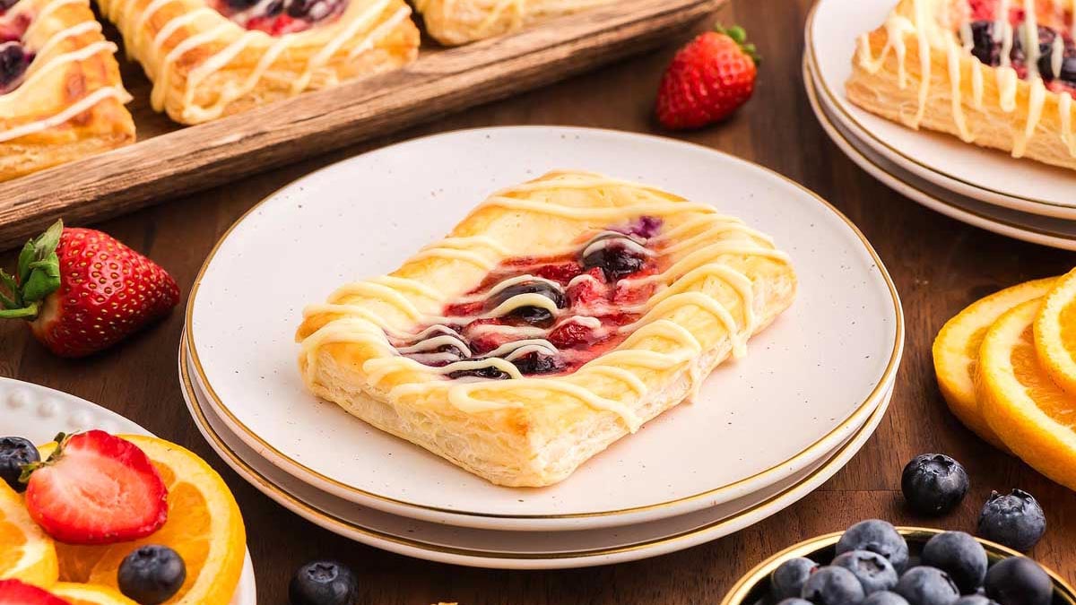 A rectangular fruit danish with icing drizzle sits on a white plate, surrounded by fresh berries and sliced oranges on a wooden table.
