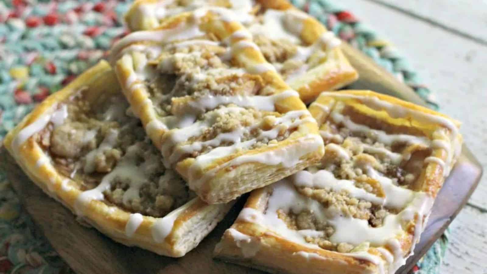 Rectangular pastries with crumb topping and white icing drizzle are stacked on a wooden board over a woven placemat.