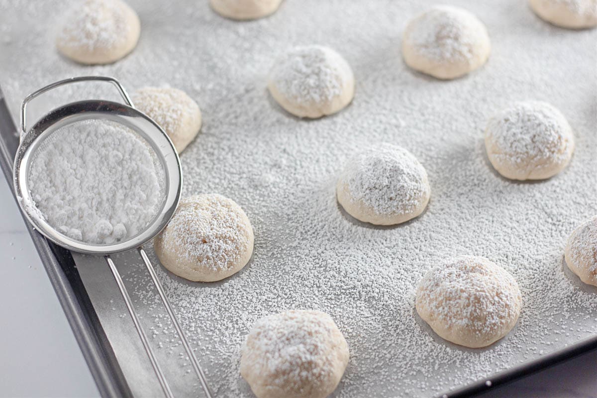 Balls of dough are arranged on a baking sheet lined with parchment paper, dusted with powdered sugar. A fine mesh strainer with powdered sugar sits on the tray.