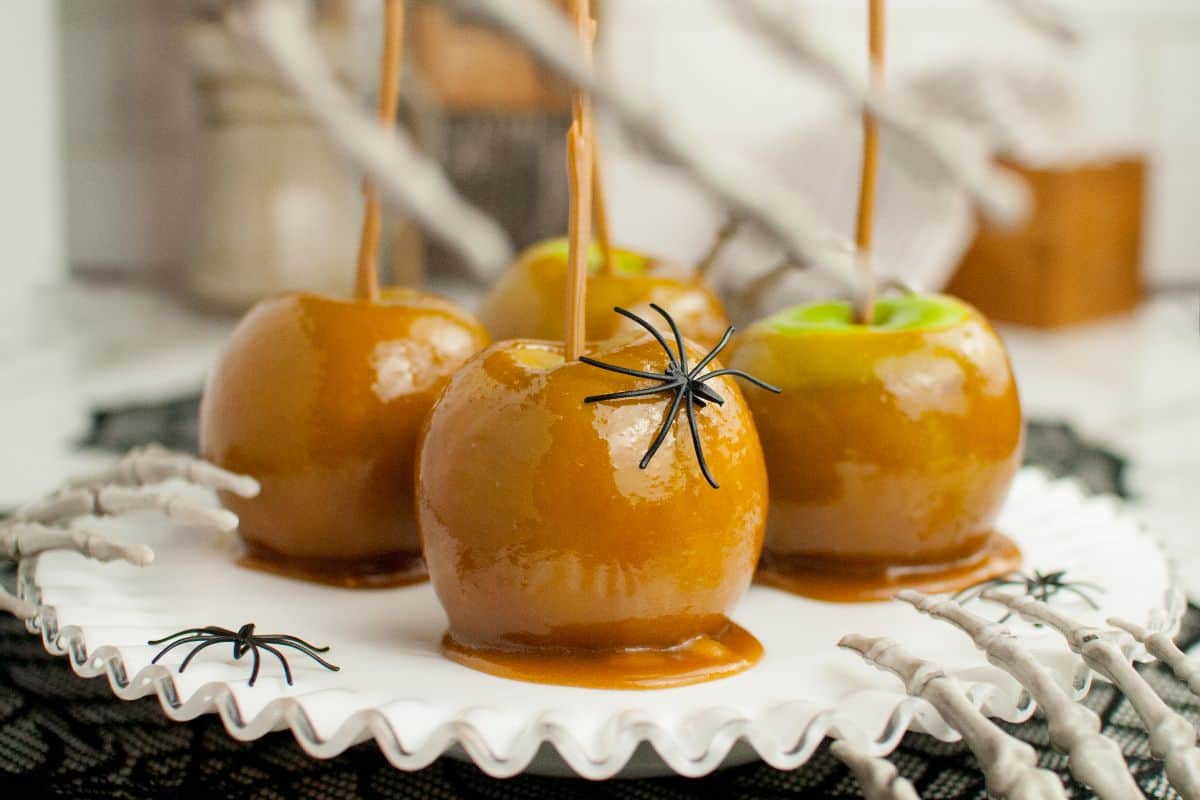 Caramel apples on a white plate, decorated with plastic spiders and skeletal hands, set on a black lace cloth.